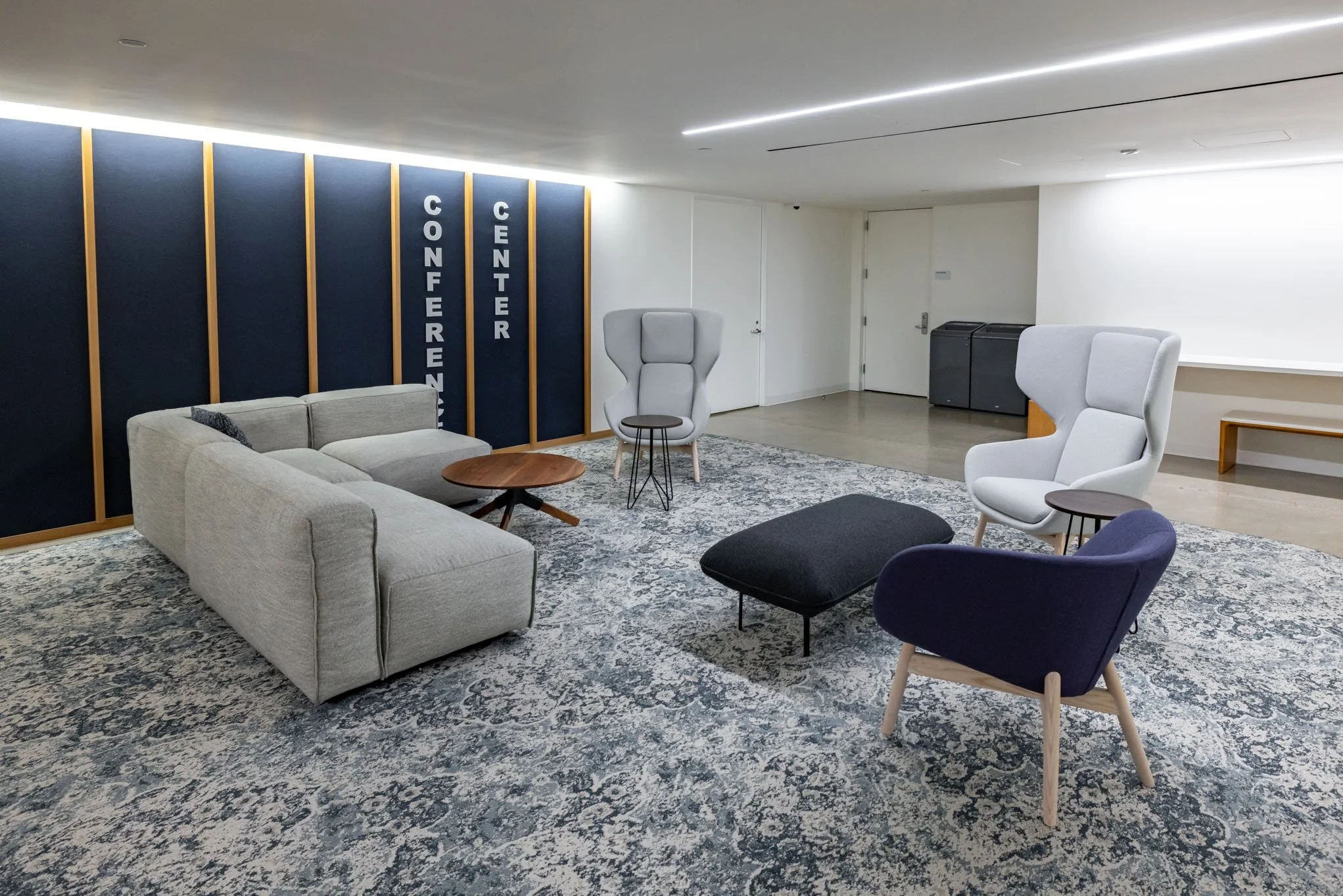 Modern conference room with gray sofa, white and navy blue armchairs, small round tables, navy and gray patterned rug, and a dark blue wall with white lettering that reads "Conference Center."