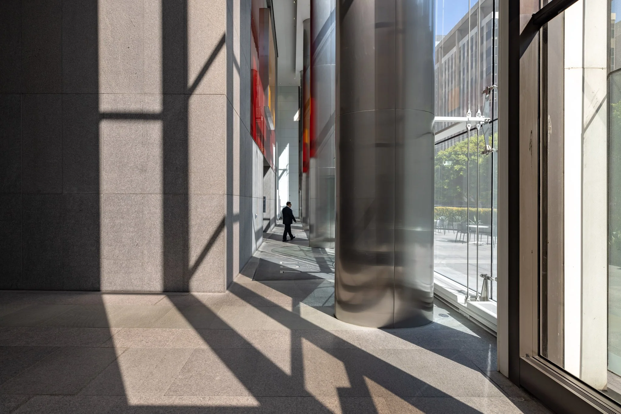 Interior of a modern commercial building with large glass windows. Sunlight creates shadows on the floor and wall, a man in business attire walks past a metallic cylindrical column.