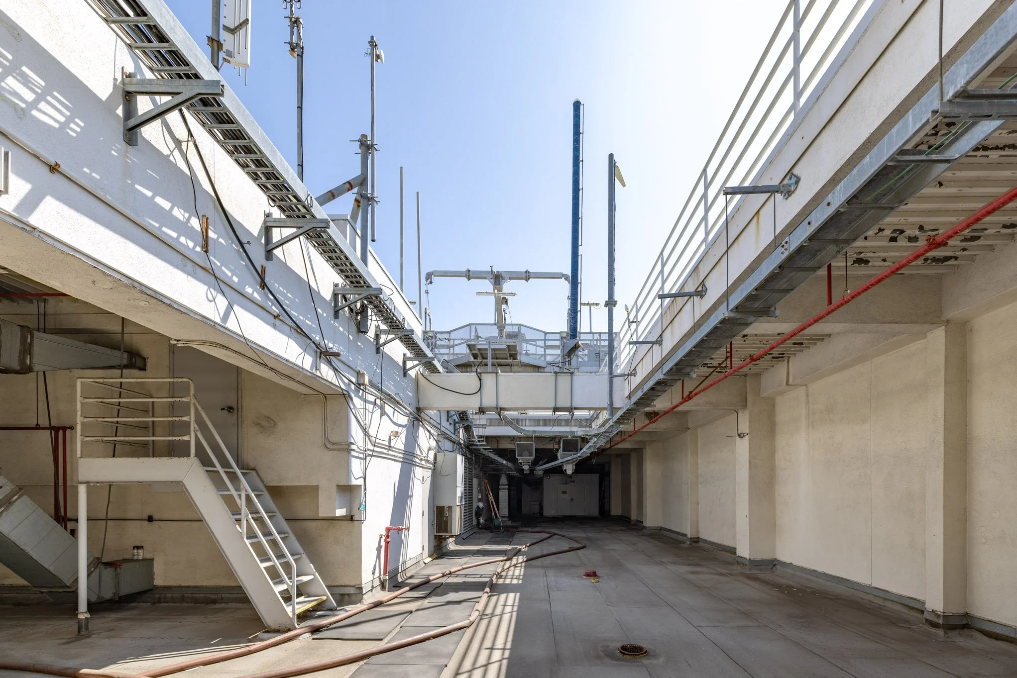 Empty rooftop terrace with electrical and plumbing infrastructure, clear blue sky, shadows on beige walls and floor.