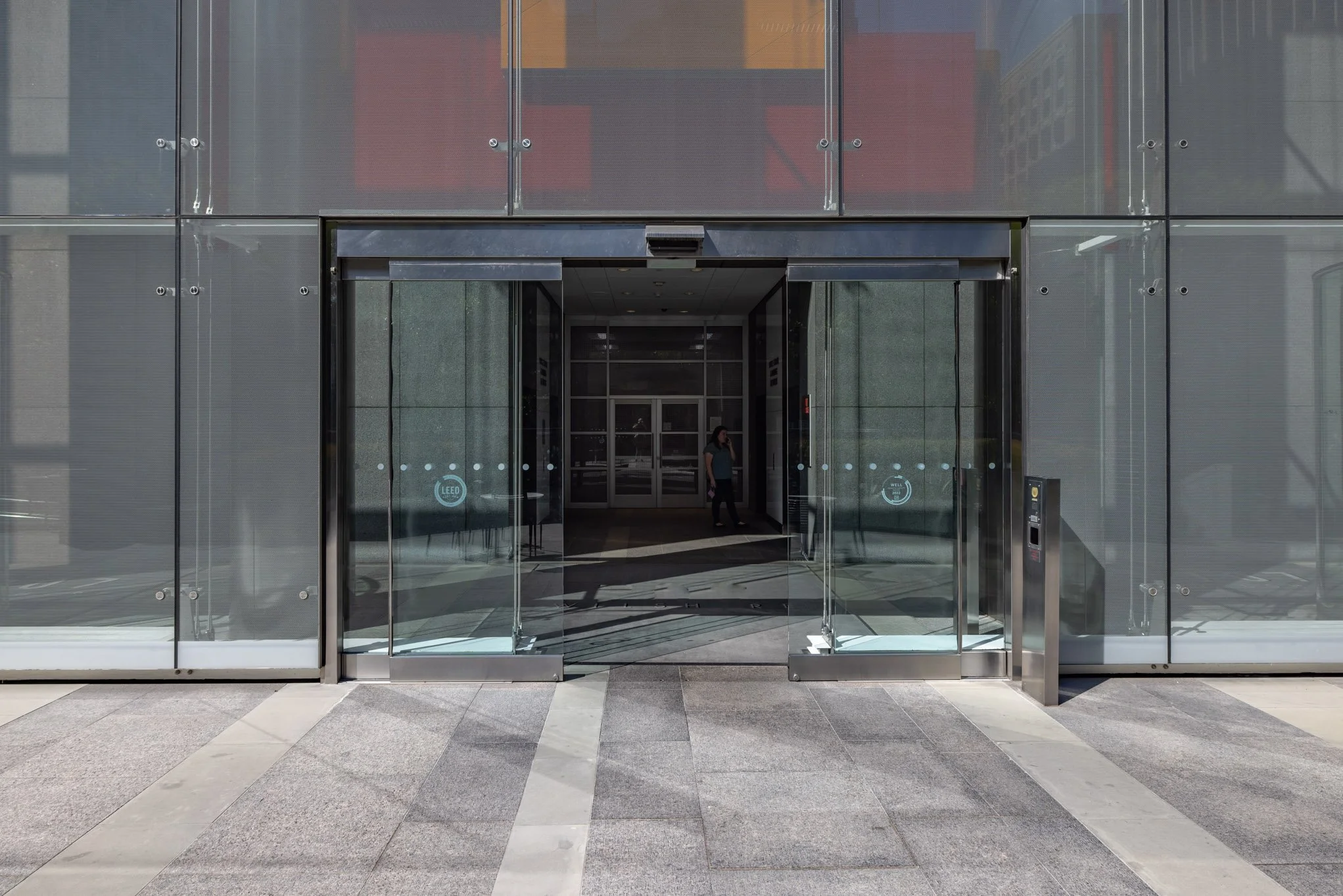 Entrance to a modern office building with glass doors and reflection of a woman walking inside.