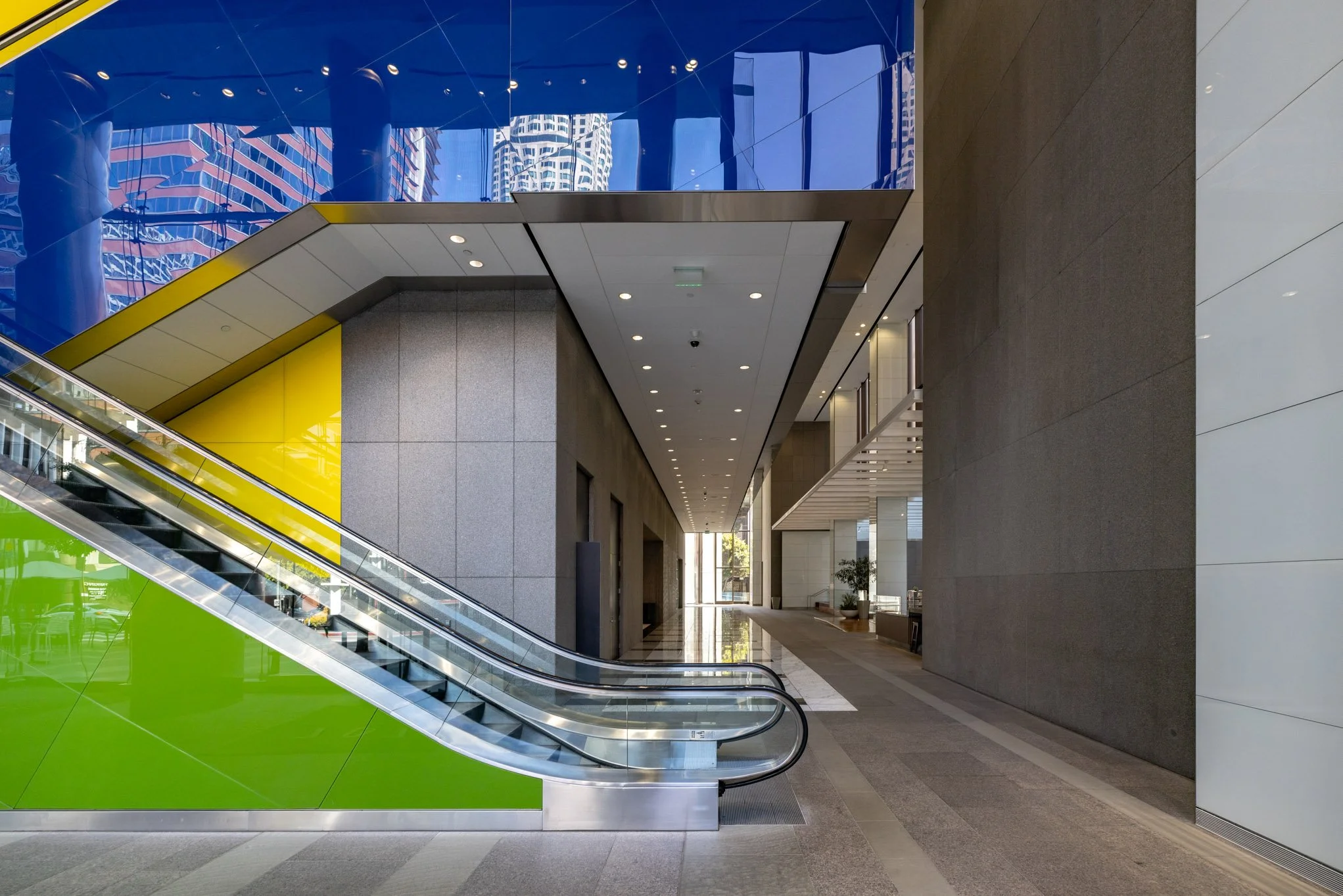 Modern office building lobby with escalator, glass walls, and a corridor leading to a glass door.