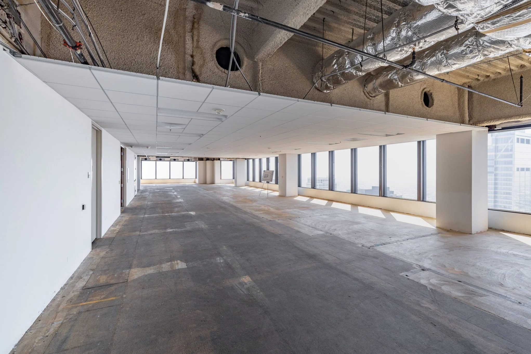 Empty office space under renovation with exposed ceiling and large windows showing city skyline.