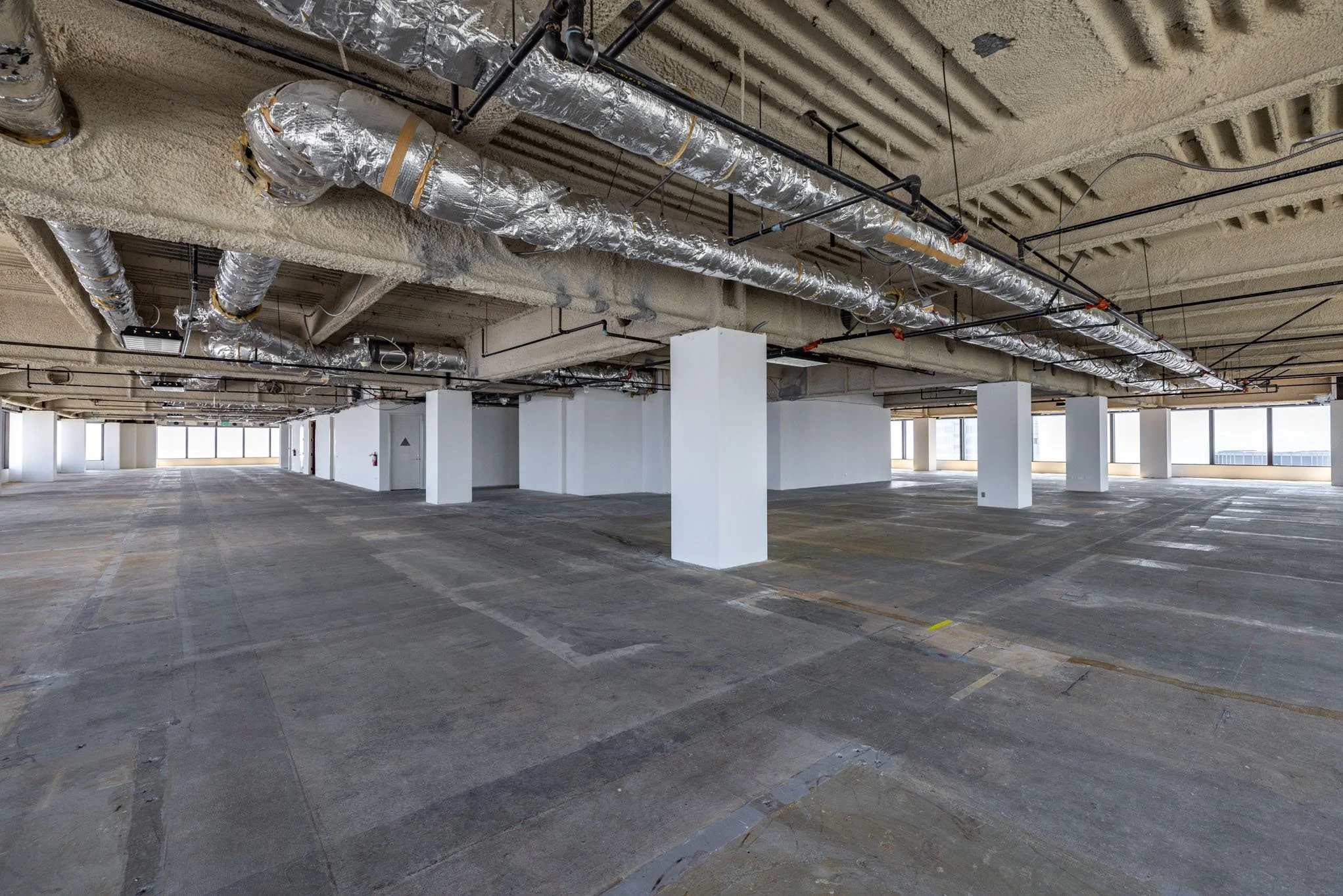 Empty indoor parking garage with concrete floors, white columns, exposed ceiling with ductwork, and large windows in the background.