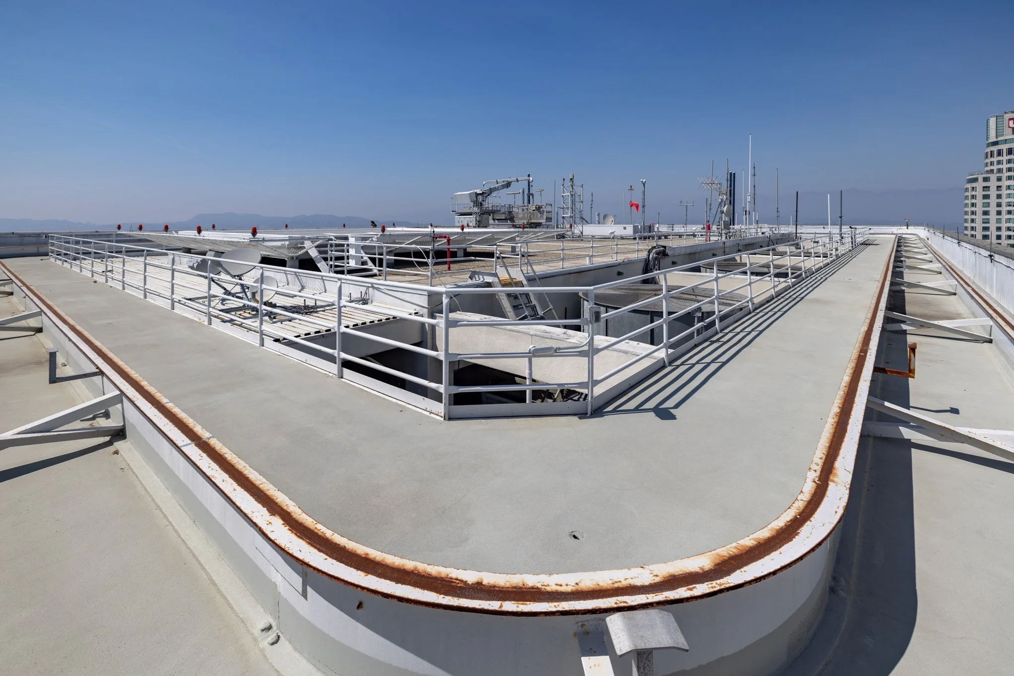 The rooftop of a building with metal railings, vents, antennas, and a clear view to the horizon, with a cityscape and distant mountains under a blue sky.