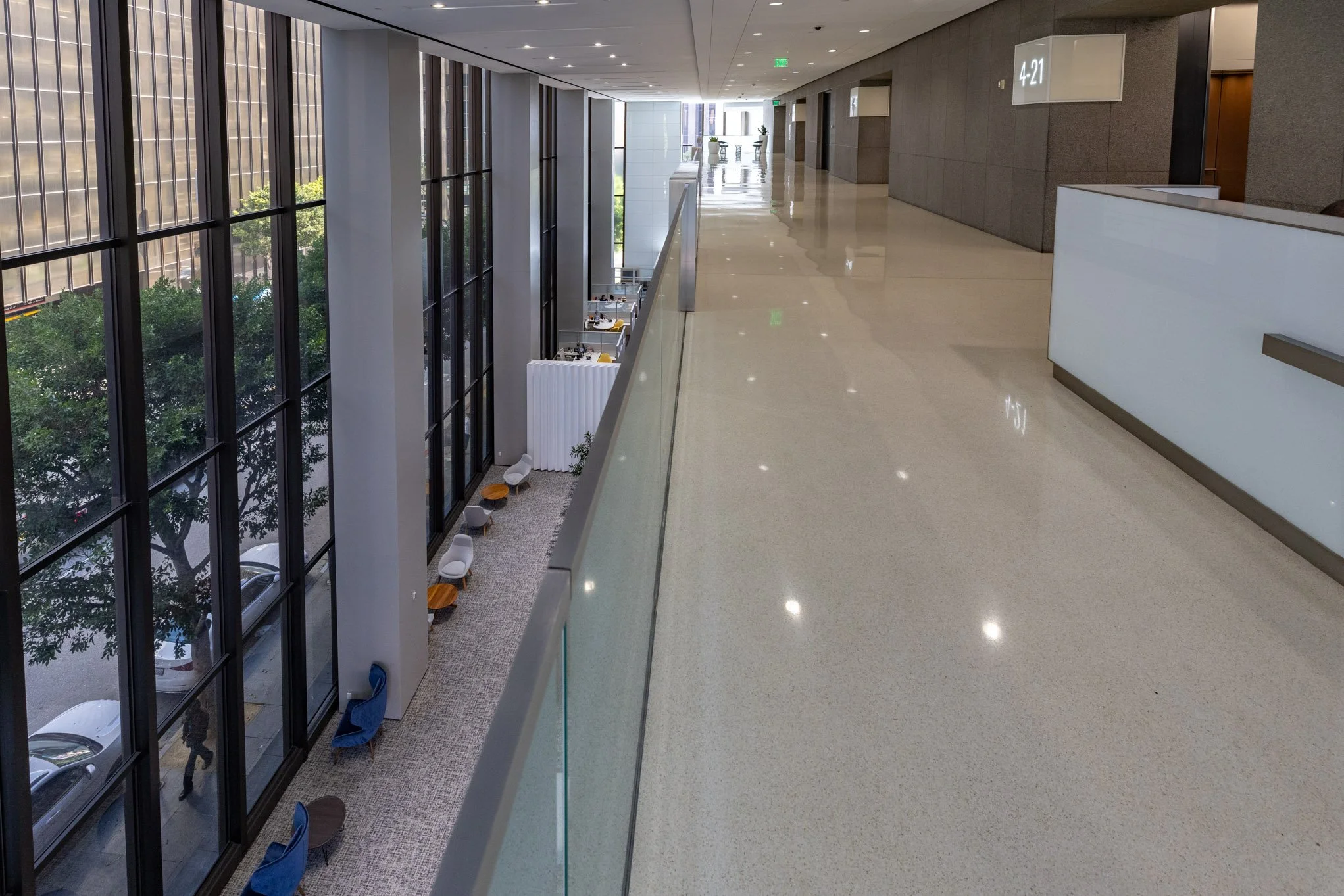 An indoor view of a modern building corridor with large glass windows on the left, overlooking a street with parked cars and trees outside. The corridor has a beige polished floor, gray walls, and embedded ceiling lights. On the right, there is a rec