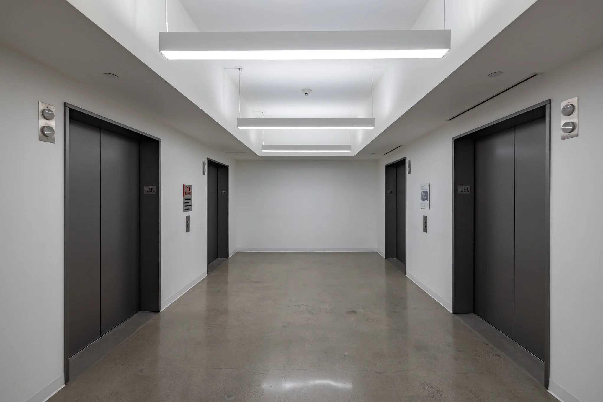 Empty elevator lobby with four closed elevator doors, white walls, polished concrete floor, and modern ceiling lights.