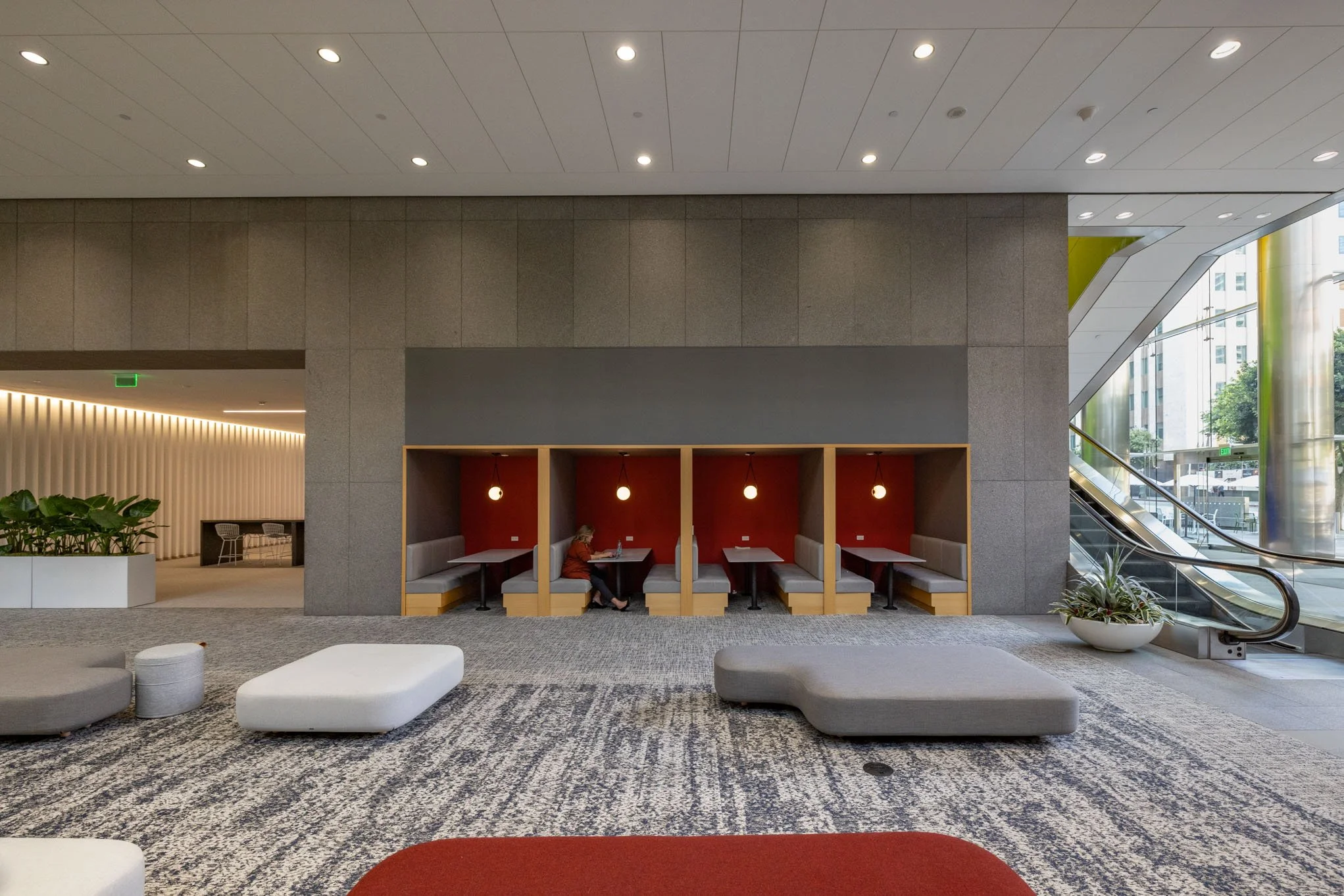 Modern lobby with seating area, including a woman using a laptop in a booth with red walls, large windows, and indoor plants.