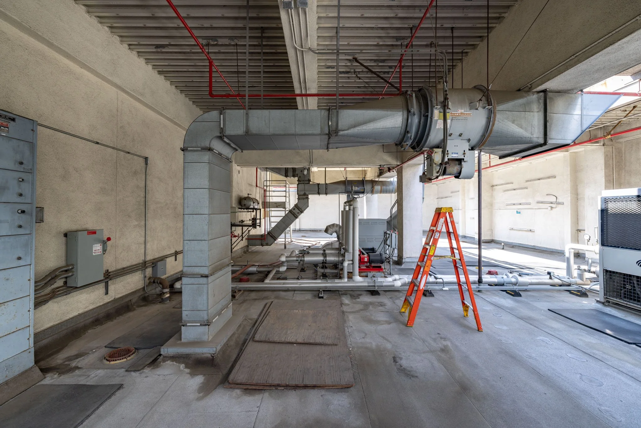 Indoor industrial area with large metal ventilation ductwork, pipes, a red ladder, and electrical boxes on the wall.