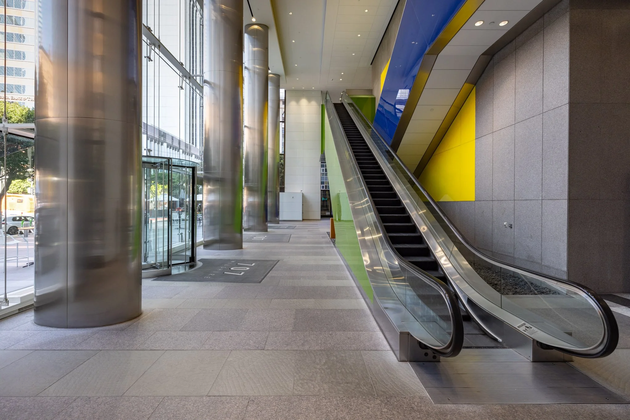 Interior of a modern building with large glass windows, shiny metallic columns, an escalator, and colorful wall panels in blue and green.