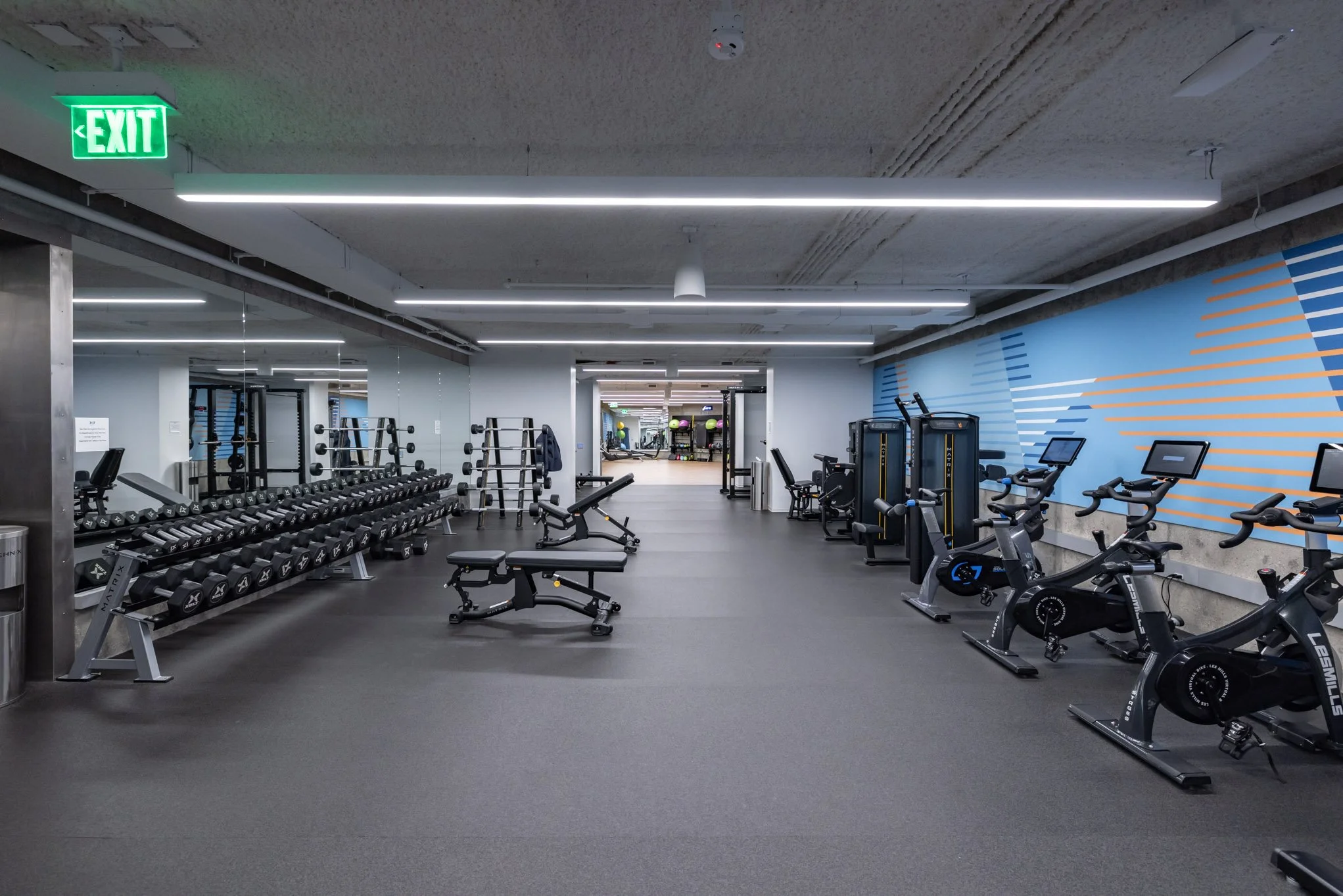 Empty gym with free weights on the left, exercise bikes on the right, and various workout machines in the background, illuminated by ceiling lights and an exit sign.