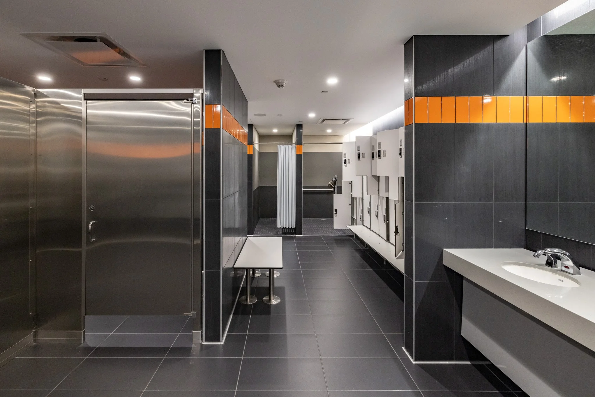 Public bathroom with black tiles, orange accents, stainless steel stalls, white lockers, a sink, and a small bench.