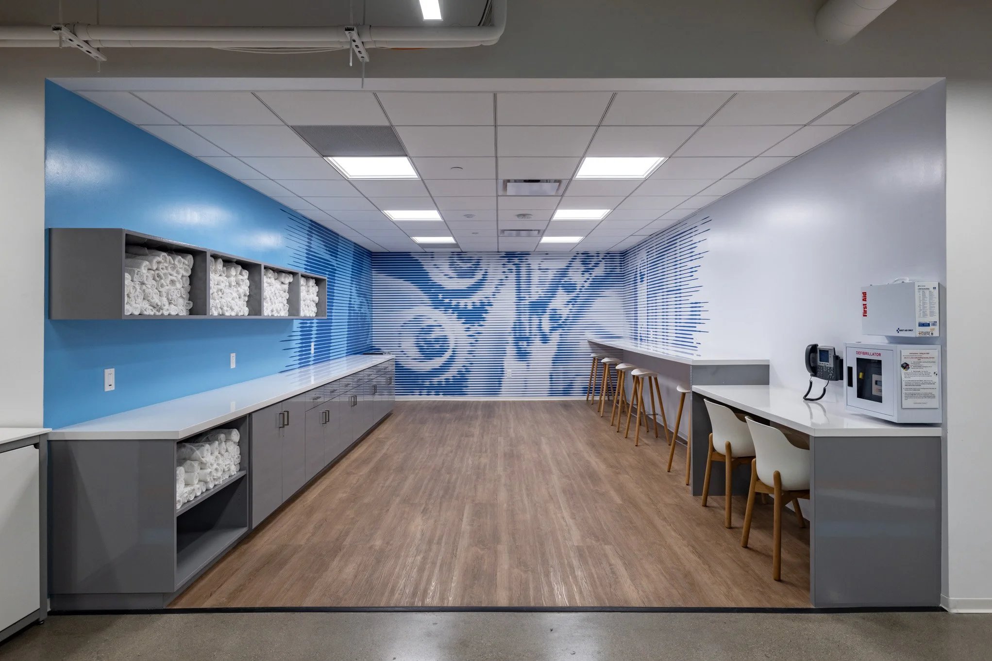 Empty lounge area with blue and white maya mural on back wall, counter with white chairs, and shelves with white towels.