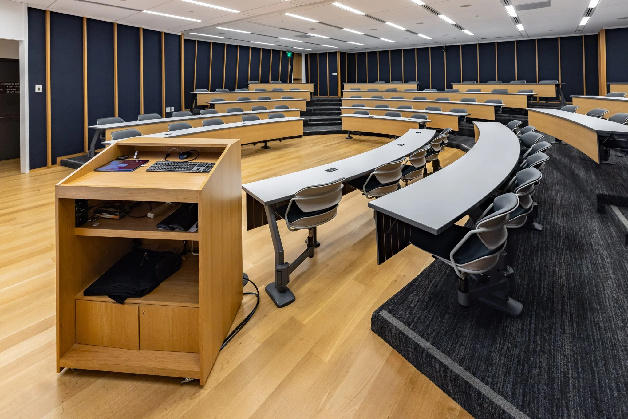 Empty university lecture hall or classroom with tiered seating, rows of chairs and desks, a wooden podium with electronic equipment, and blue and wood-paneled walls.