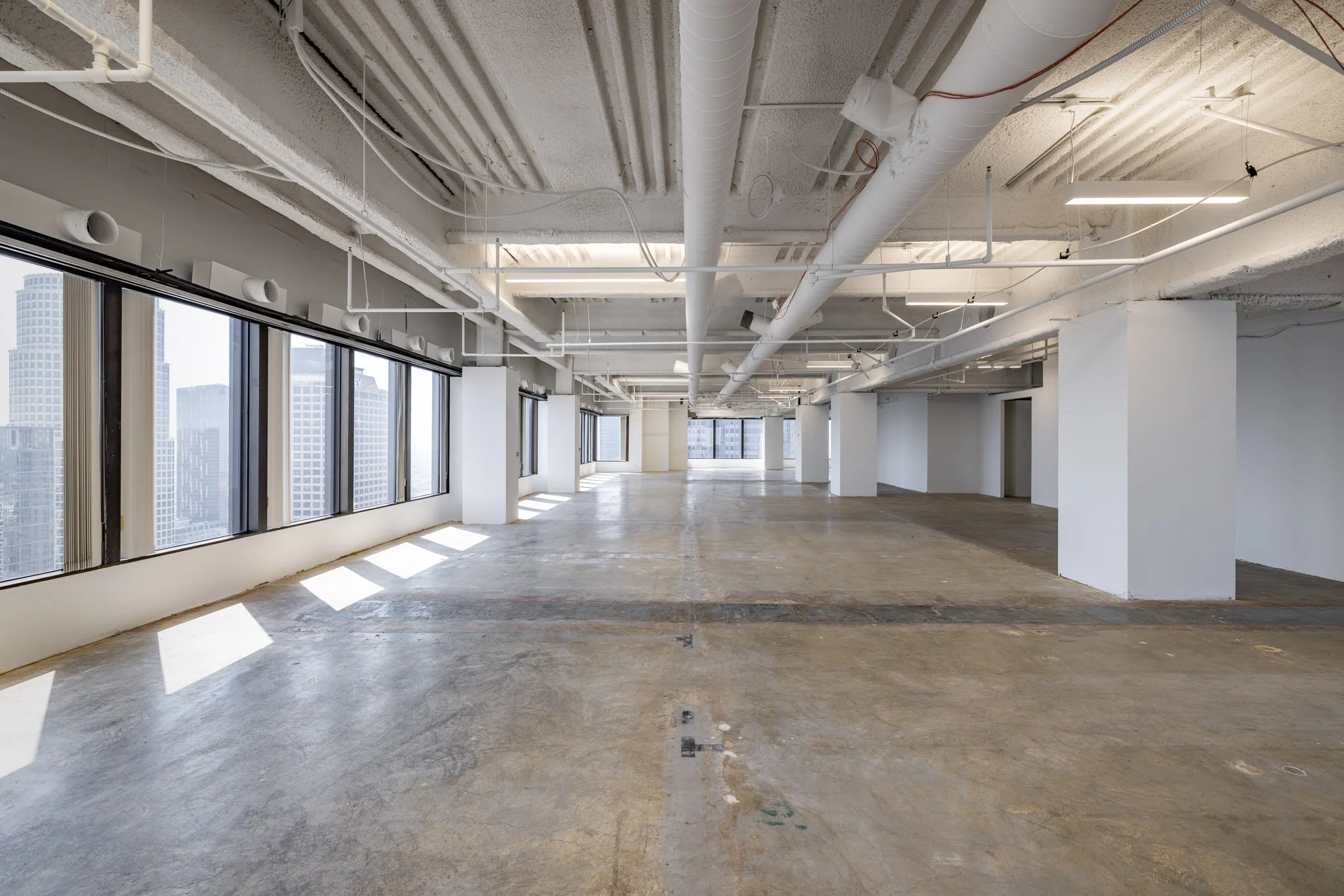 Empty commercial space with large windows, concrete floor, exposed ceiling pipes, and city view.