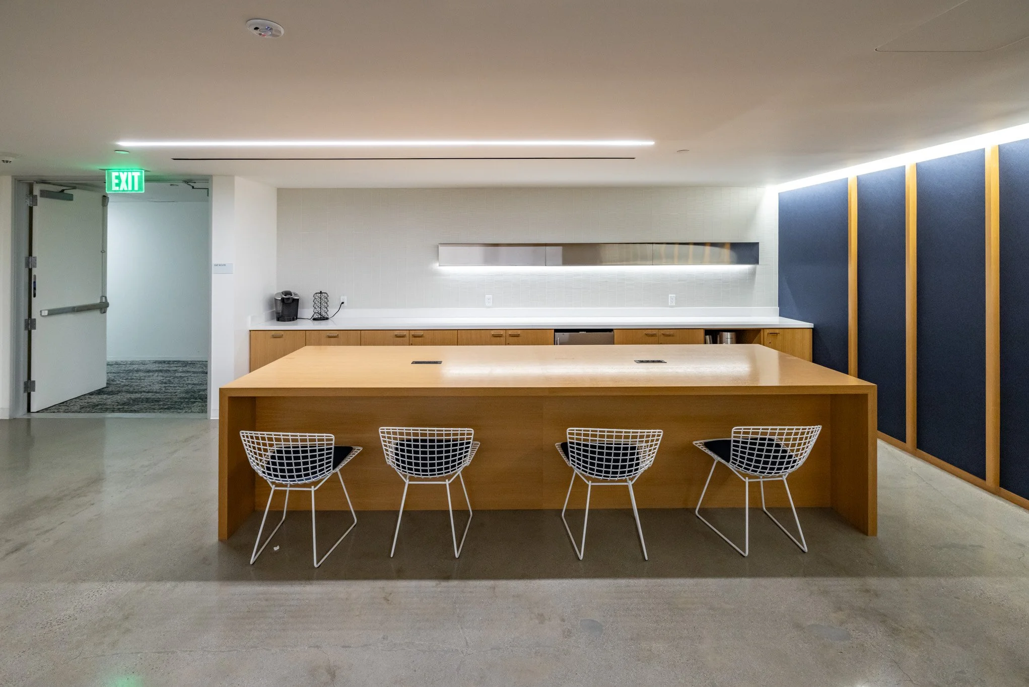 Modern kitchen with a large wooden island, four wireframe chairs, a coffee maker, and a horizontal window on the back wall.