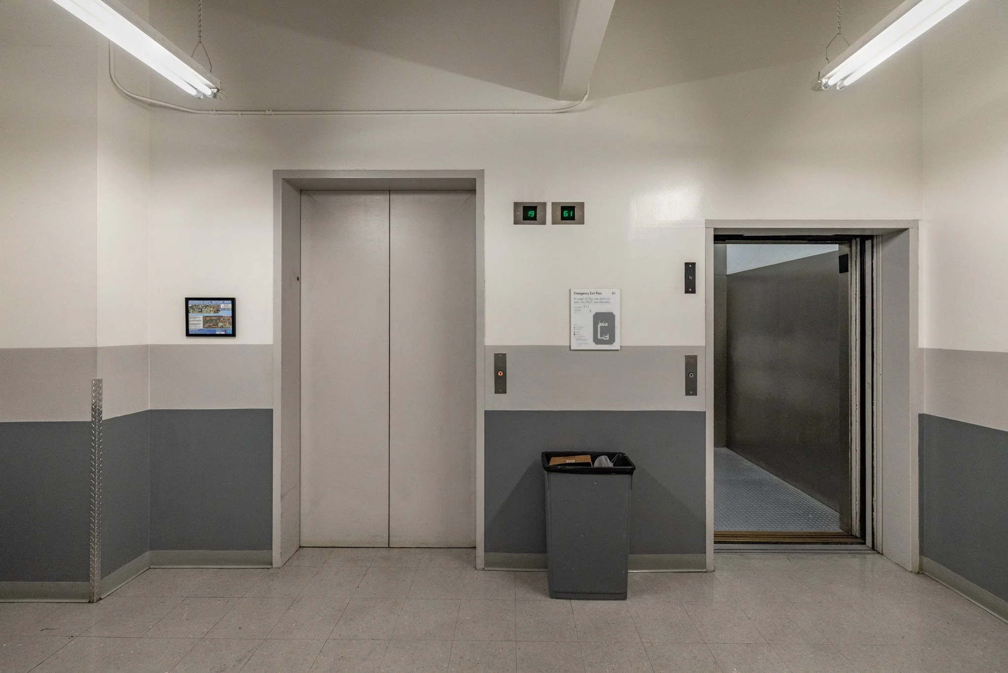 Elevator lobby with two elevator doors, digital floor indicators, a trash bin, and an emergency plan on the wall.