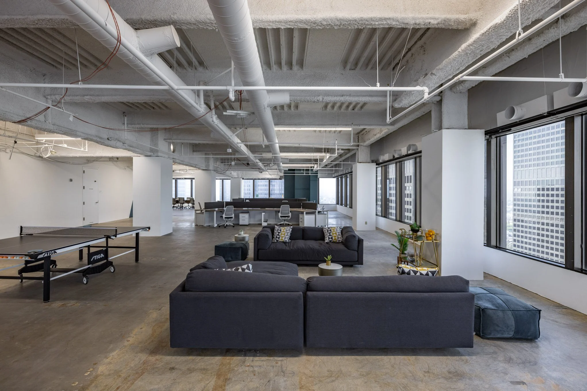 Spacious modern office with large windows, grey sofas, ping pong table, and workstations in background.