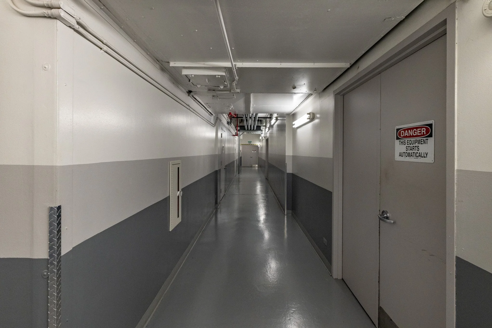 Empty industrial hallway with gray floor, white walls, and fluorescent lighting, including a warning sign on a door that says "Danger, this equipment starts automatically."