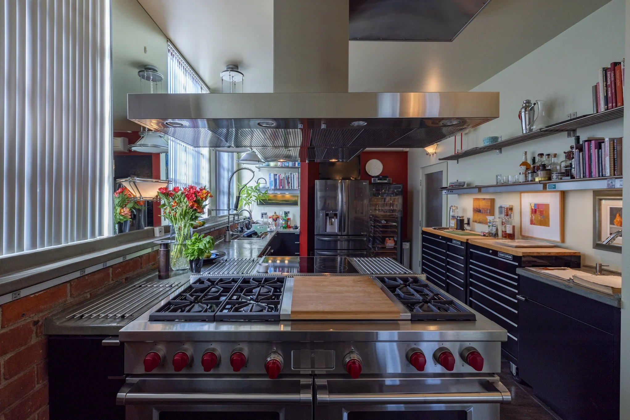 Modern kitchen with stainless steel stove, blue cabinets, open shelves with books and kitchen items, a refrigerator, and a window with vertical blinds.