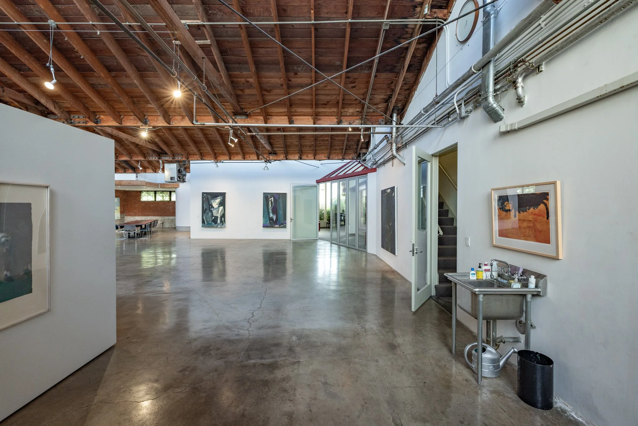 An interior view of an art gallery with white walls and wooden ceiling, displaying several abstract paintings. There's a small stainless steel sink with cleaning supplies and a black trash can near the right wall, and a door with glass panels leading