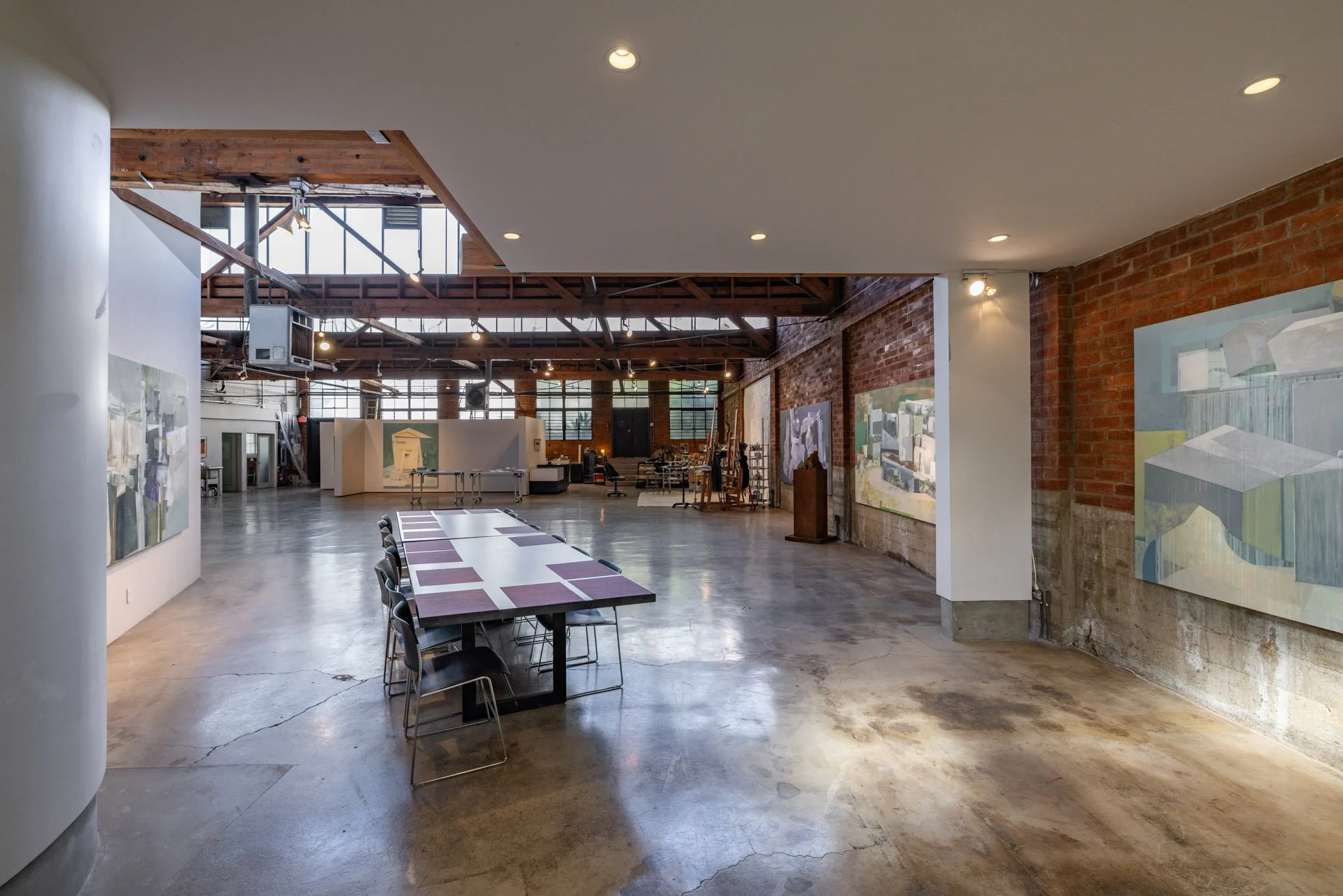 Art gallery with paintings on the walls, a long table with chairs, and various art supplies and furniture in the background.