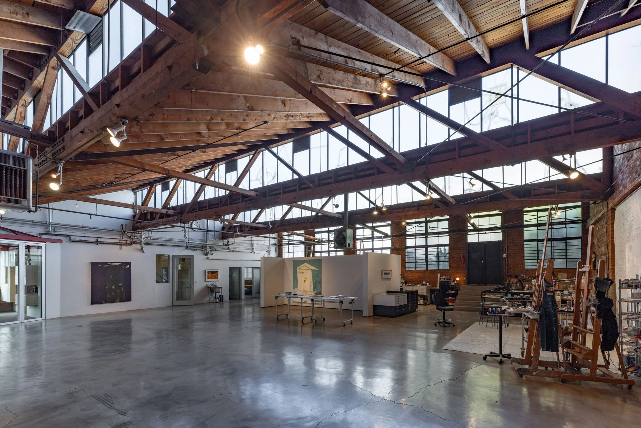 Spacious art gallery with high ceiling, large windows, wooden beams, and artworks on white walls, with art supplies and easels on the right side.