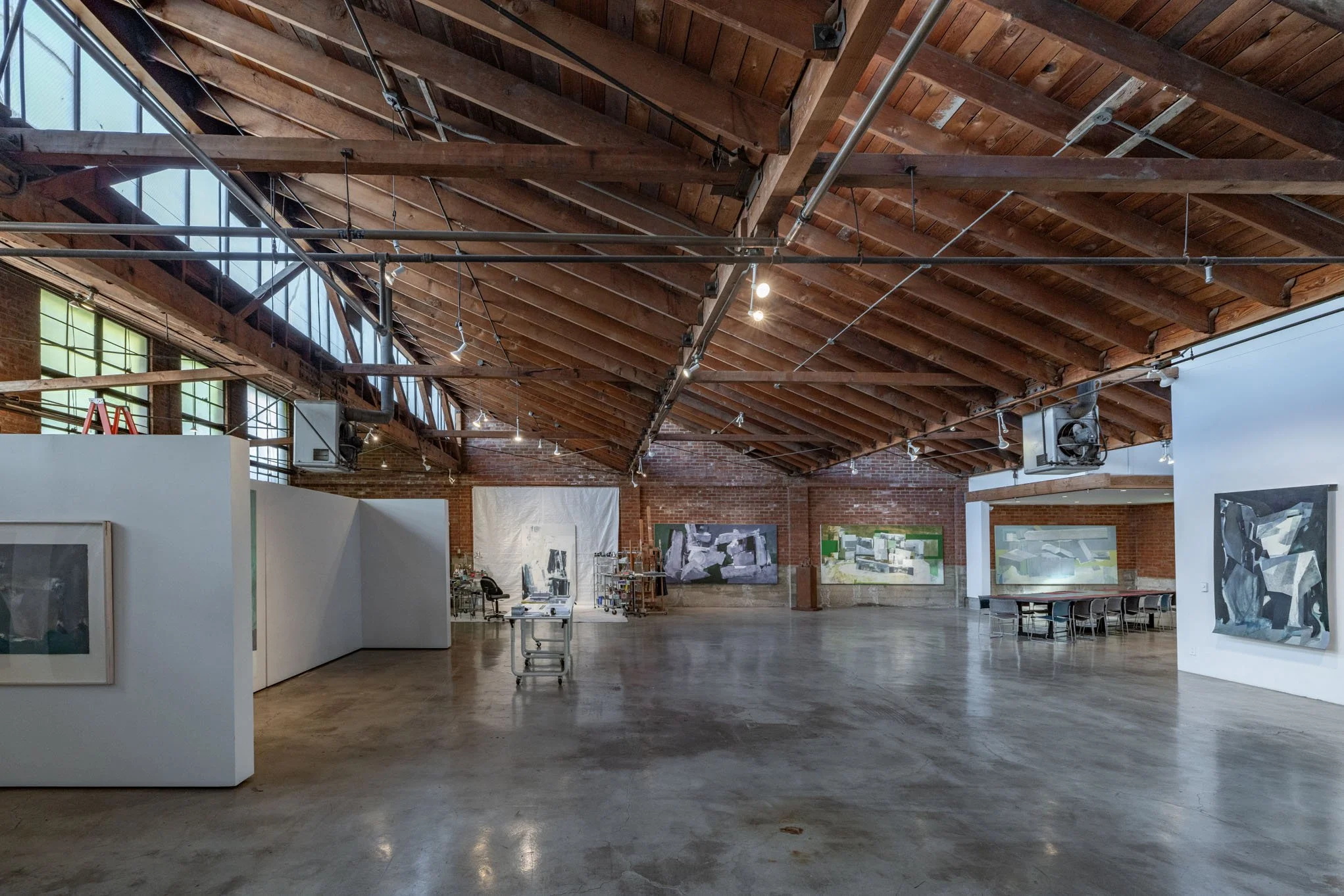 An art gallery with paintings displayed on white walls and exposed brick walls, high wooden beam ceiling, polished concrete floor, and large windows letting in natural light.