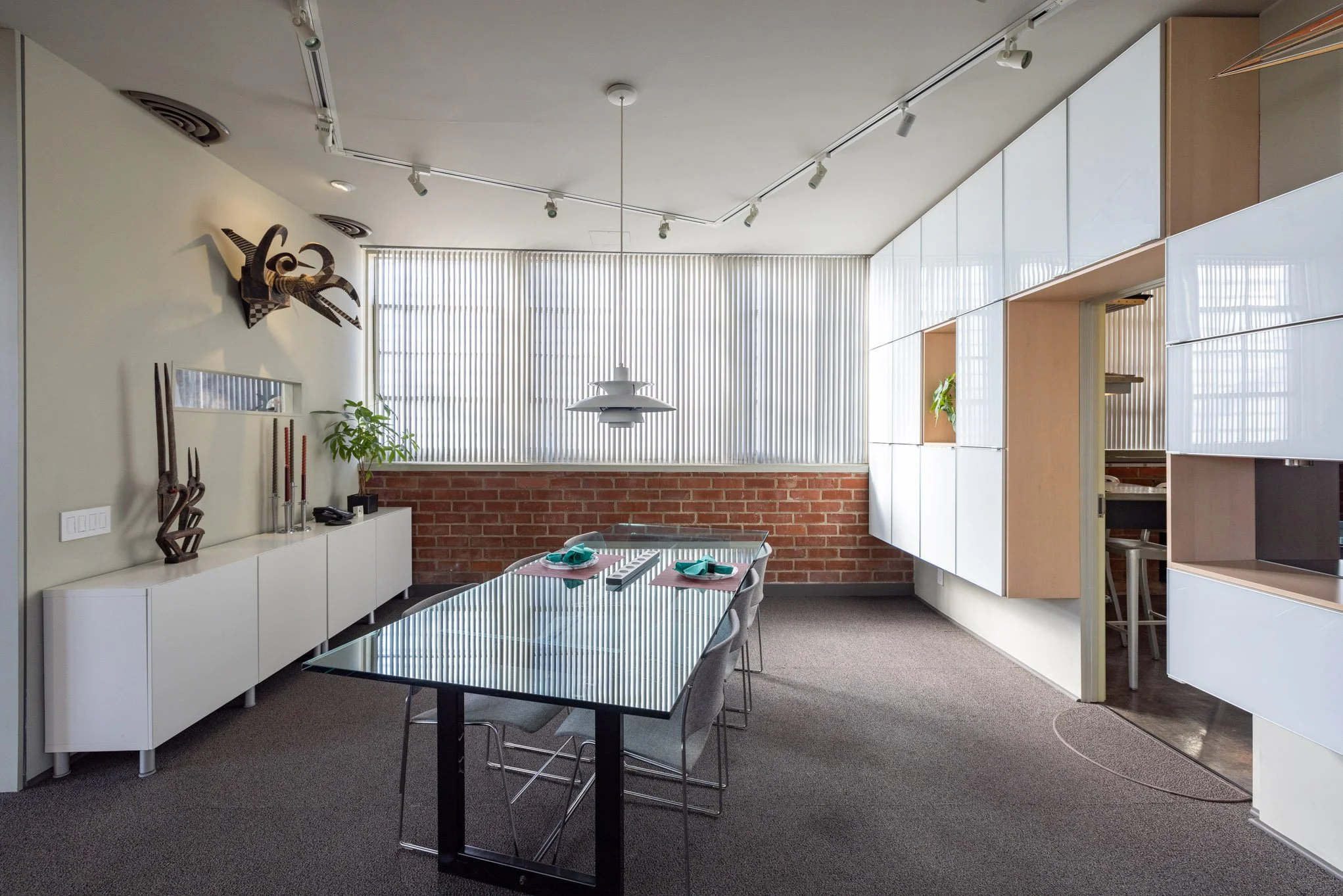 Modern dining room with a glass table, gray chairs, a white sideboard, and decorative sculptures, brick wall, vertical window blinds, and built-in cabinets.
