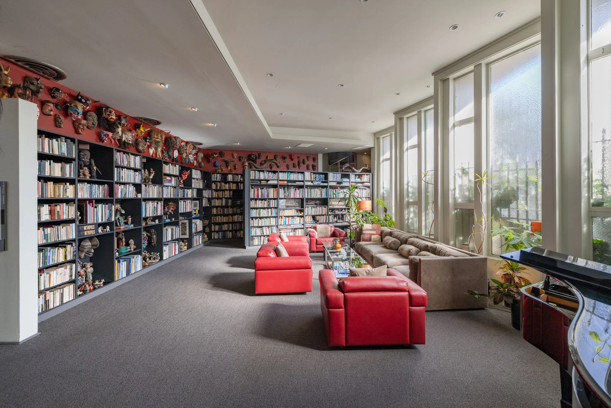 A spacious room with large windows, bookshelves filled with books, and seating including red armchairs, a beige sectional sofa, and potted plants, with decorative masks and art on the walls.