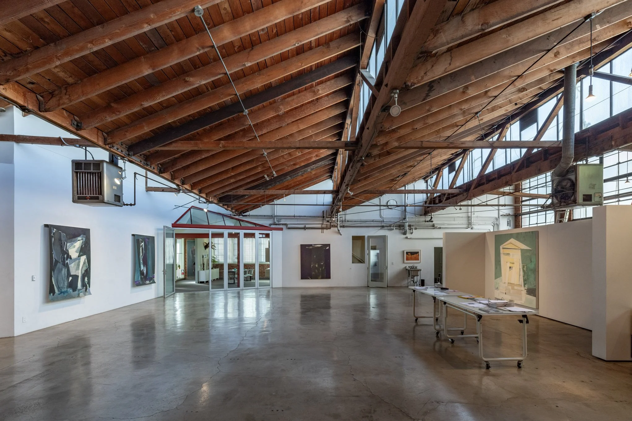 An art gallery with paintings displayed on white walls, a table with art materials, and a glass-enclosed room in the background. The gallery has an exposed wooden ceiling and concrete floor.