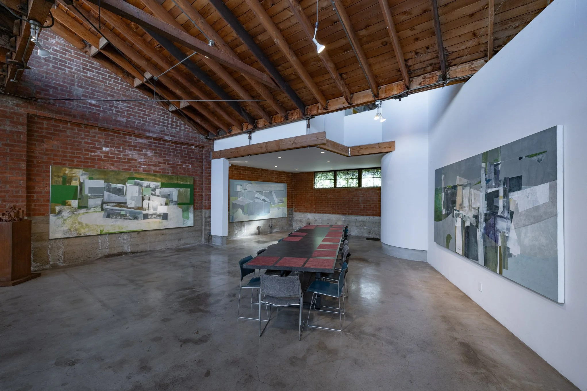 Art gallery interior with abstract paintings on brick and white walls, a long table with chairs, exposed wooden beams, and concrete floor.