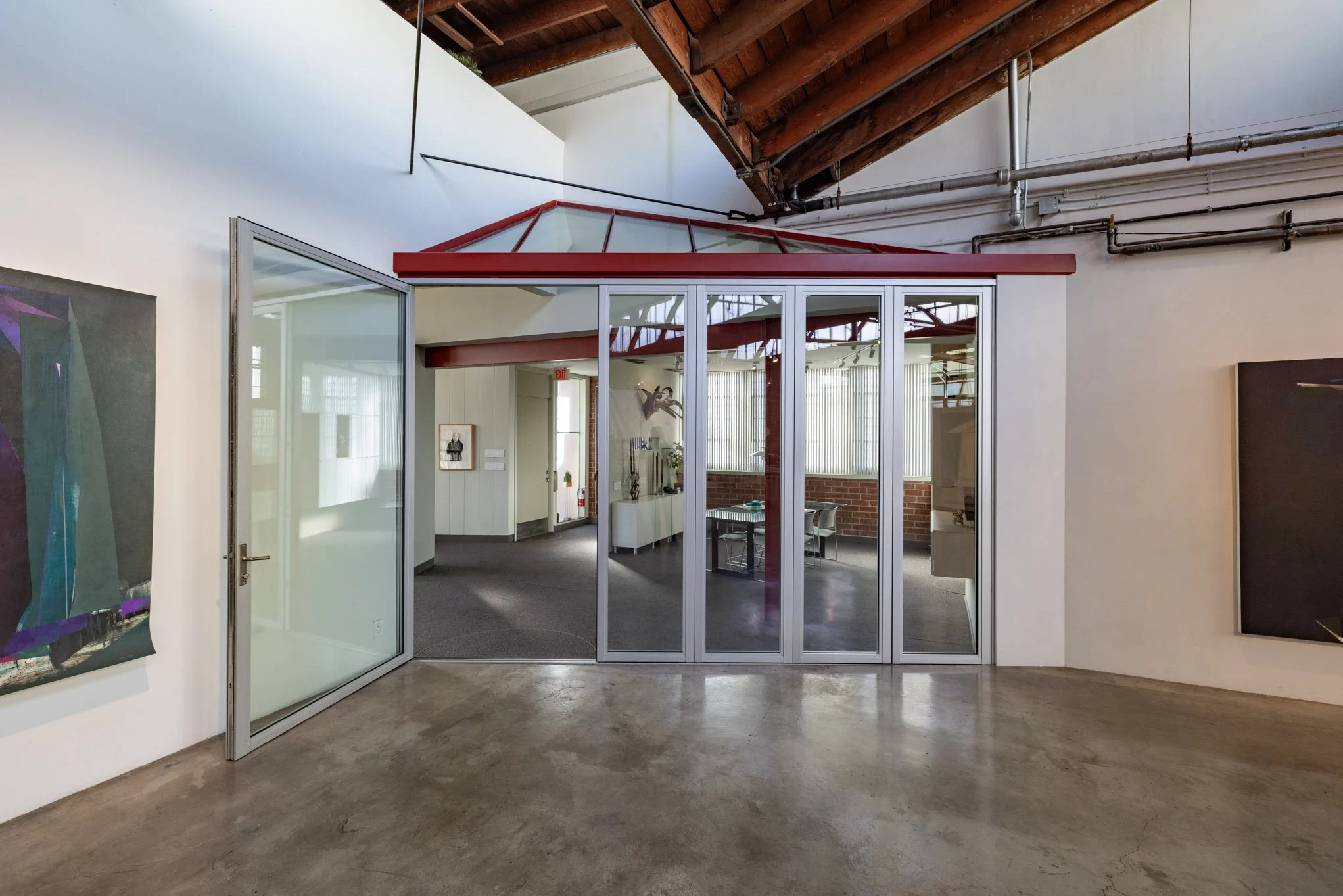 Interior of an art gallery with a glass house structure featuring red framing and a corner door, artworks hanging on the walls, and exposed wooden ceiling beams.