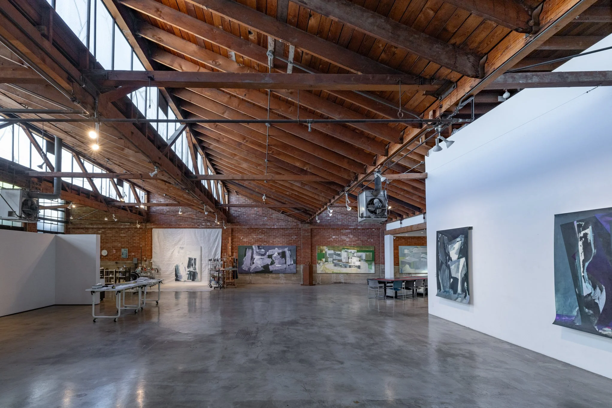 Interior of an art gallery with wooden ceiling beams, brick walls, and abstract paintings on white and brick walls, polished concrete floor, and some tables and art supplies in the background.