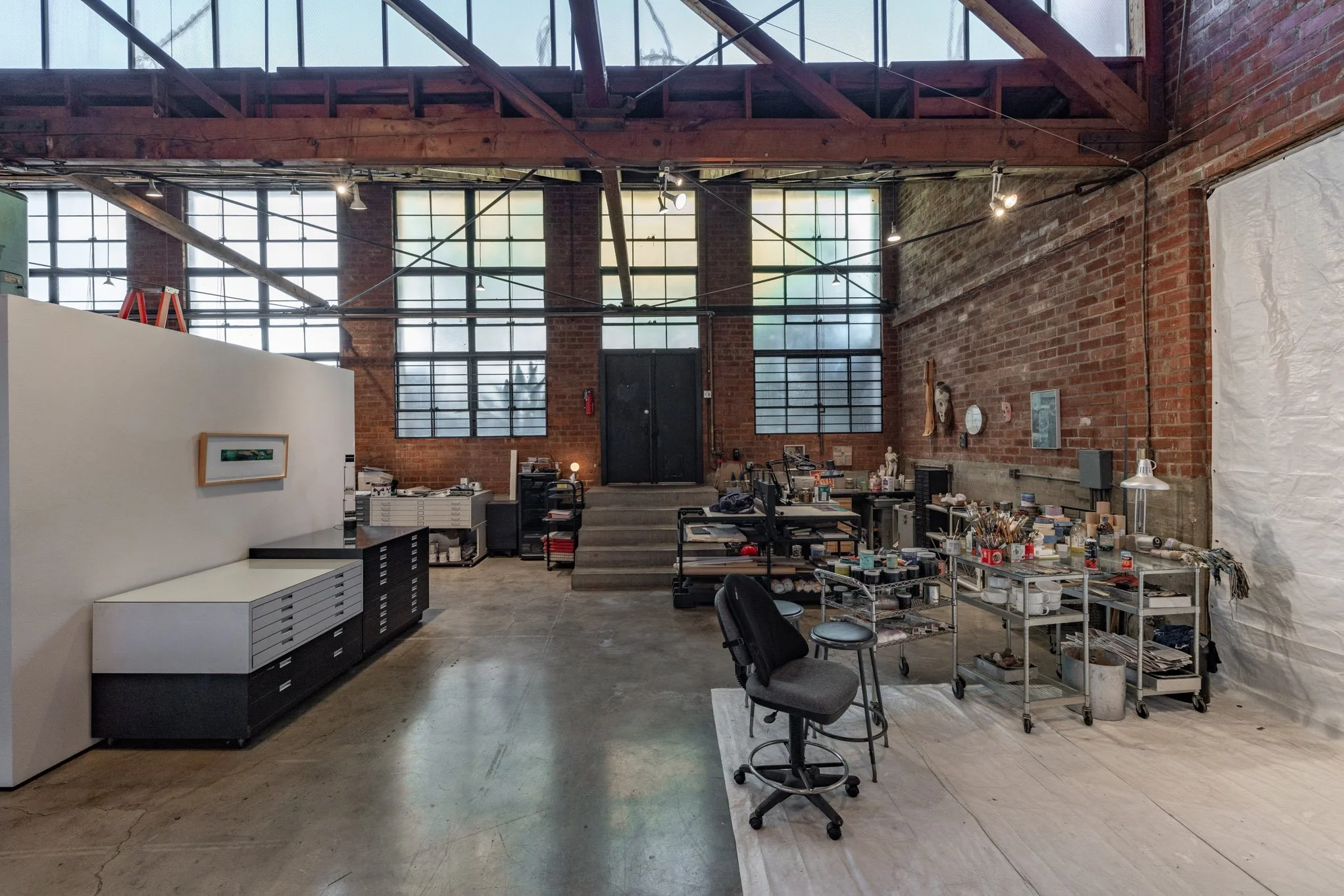 An artist's studio with brick walls, large windows, and various art supplies and tools on metal carts and tables. There are chairs, shelves, and artwork on the walls.