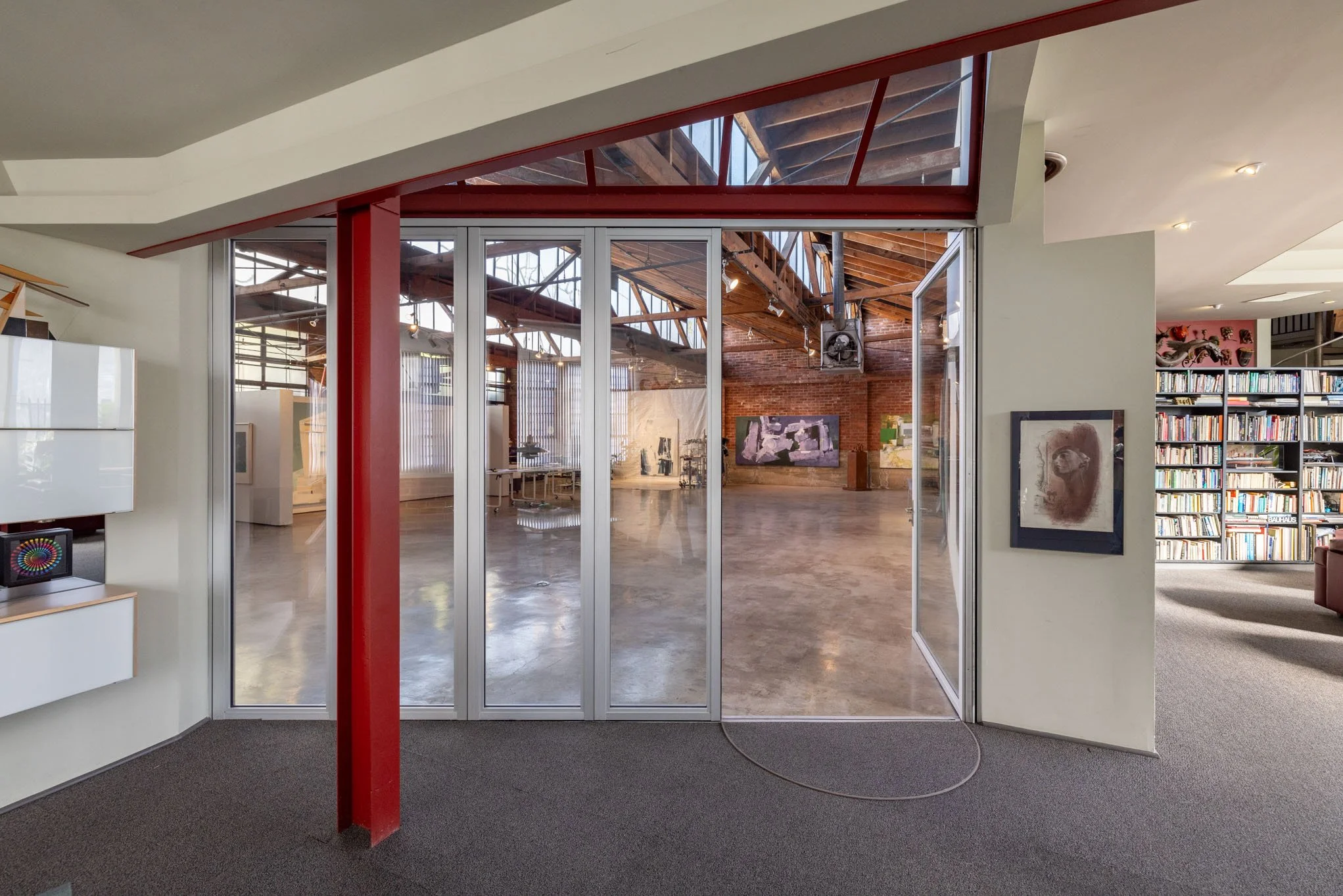 Interior view of an art gallery with large glass doors leading to a spacious room with artwork on the walls and a brick wall, metal beams, and a high ceiling.
