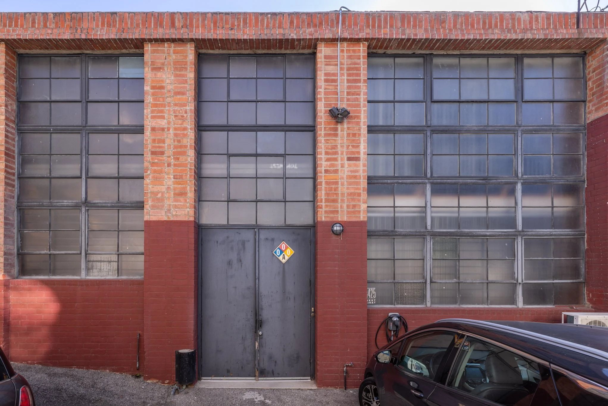 Exterior of an industrial building with large glass block windows, a black metal door with a hazard diamond sign, and a car parked in front.