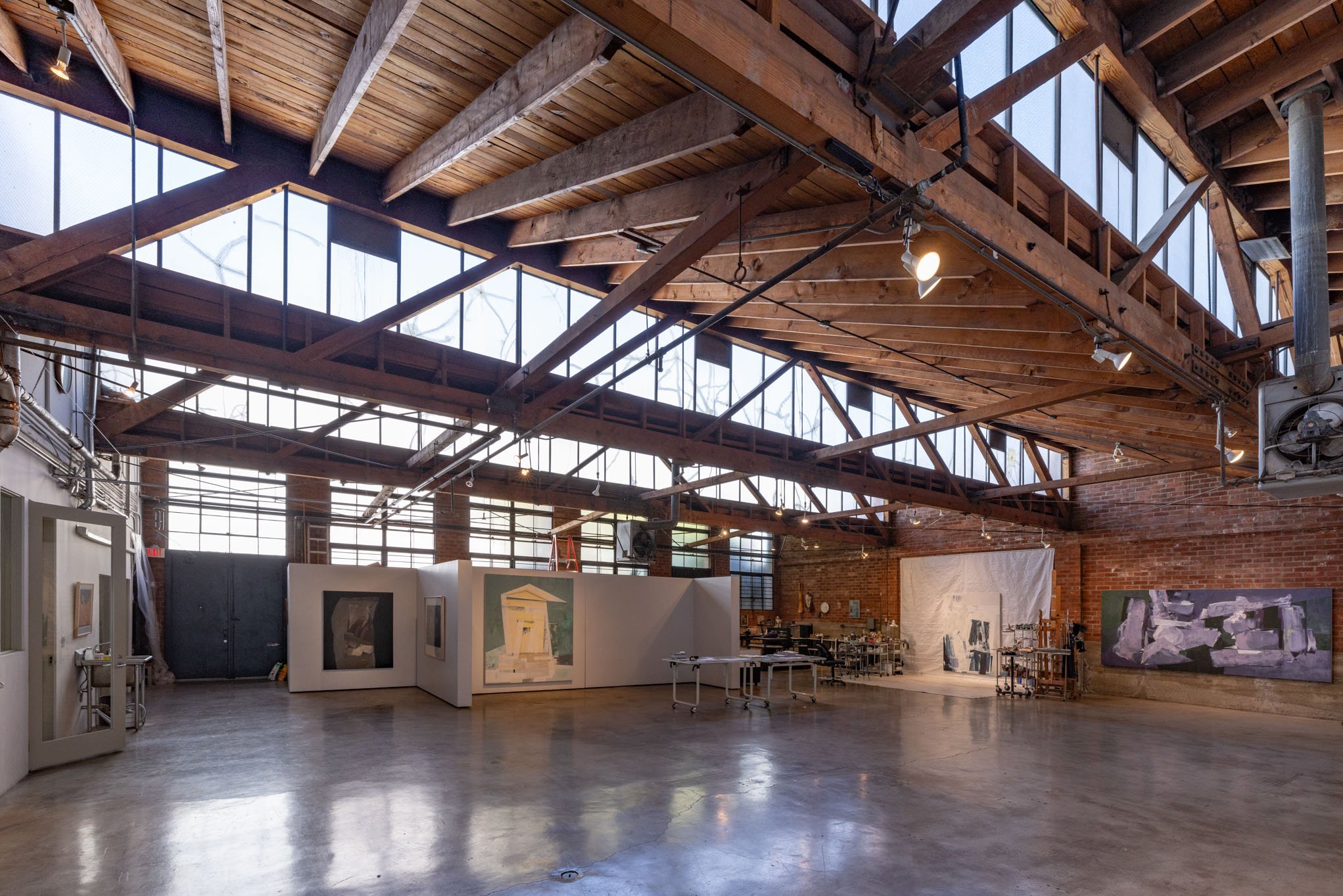 An industrial-style art gallery with exposed wooden beams and large windows, displaying various paintings and artwork on white partitions and brick walls.