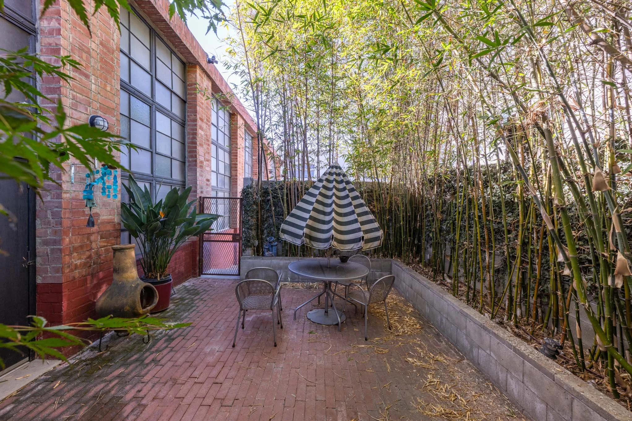 Small outdoor patio area with brick flooring, a round glass table with four metal chairs, a striped black-and-white patio umbrella, tall bamboo plants on the right, a brick wall building on the left side, and a black metal gate in the background.