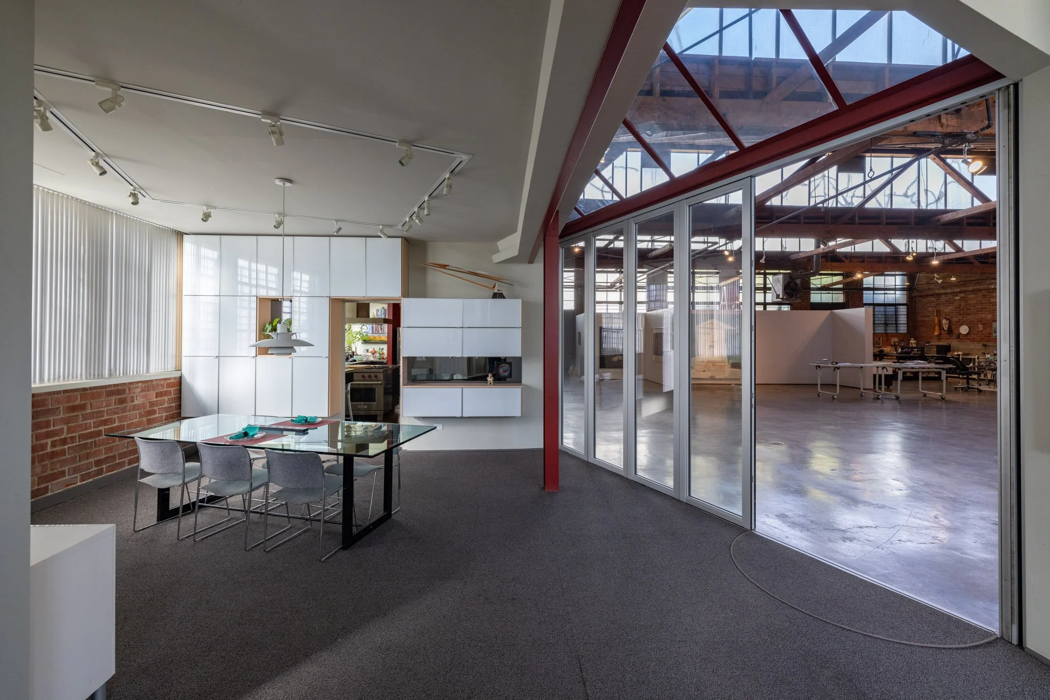 Modern open-plan office with a glass dining table set for four, white cabinetry, a brick wall, large windows, and an industrial-style conference area separated by glass walls.