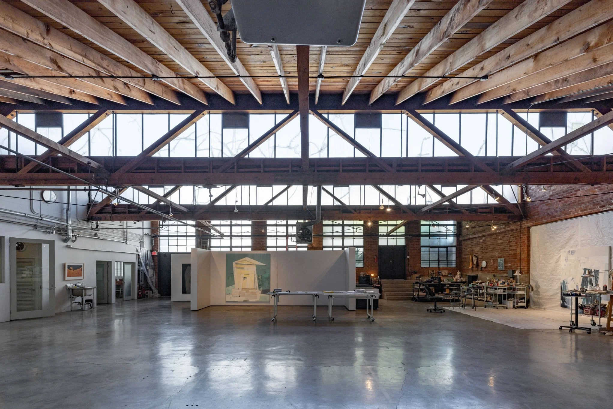 Interior of an art studio or gallery with a high ceiling, exposed wooden beams, large windows, and various artworks and workspaces.