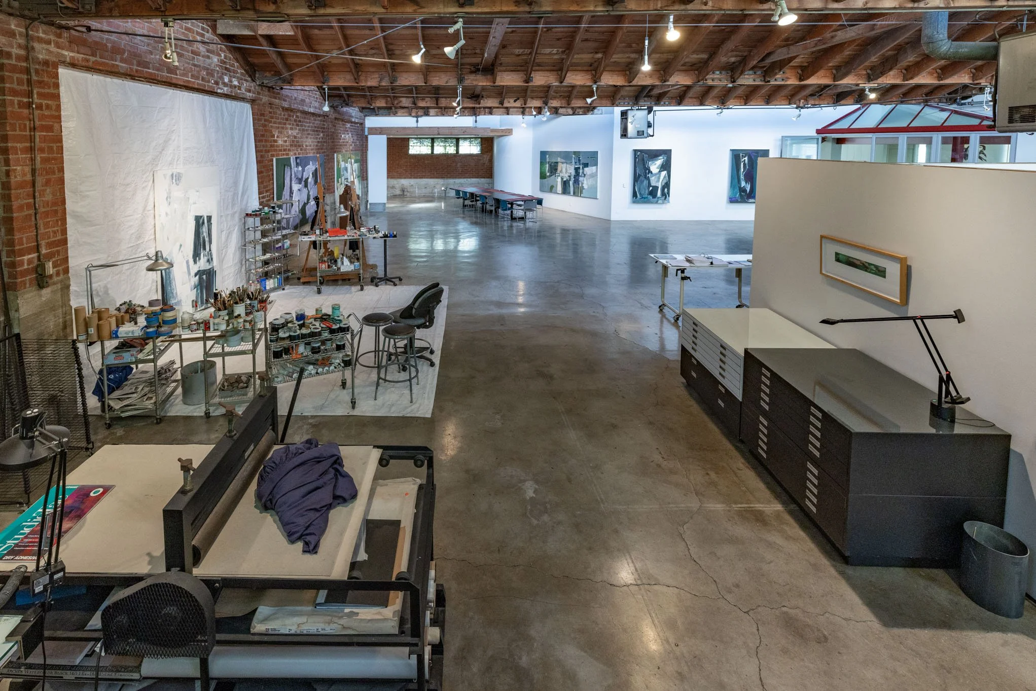 An art gallery with paintings on white and brick walls, several tables with art supplies, and a workspace with a lights and framed artwork.