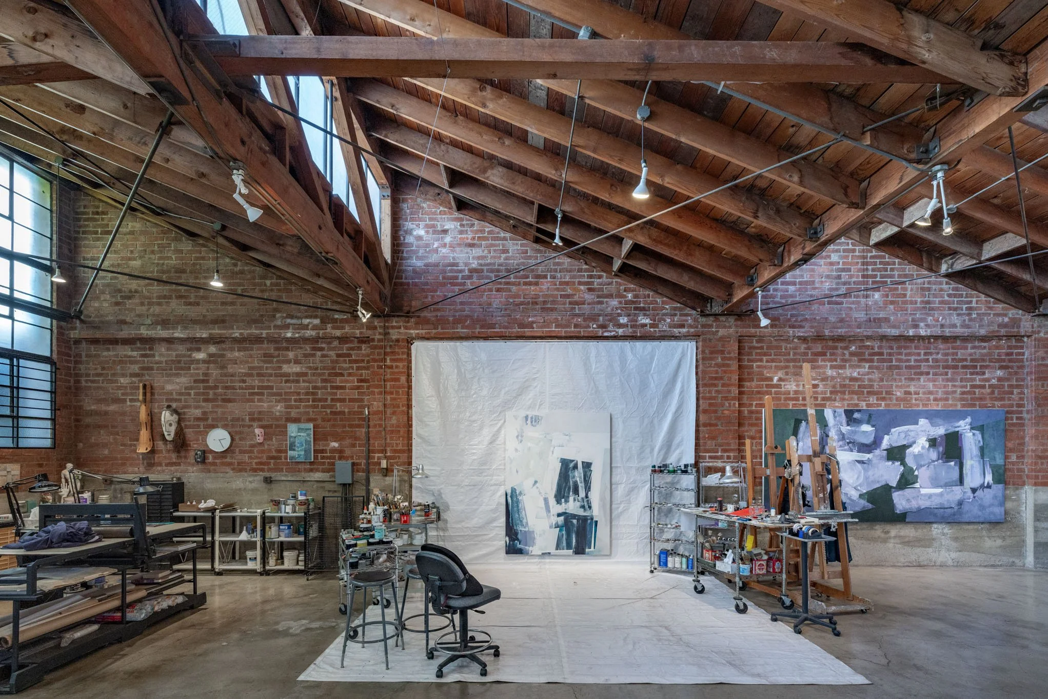 An artist's studio with a brick wall interior, wooden ceiling beams, and large windows. Art supplies, easels, and paintings are arranged throughout the space, with a white backdrop and chairs in the center.