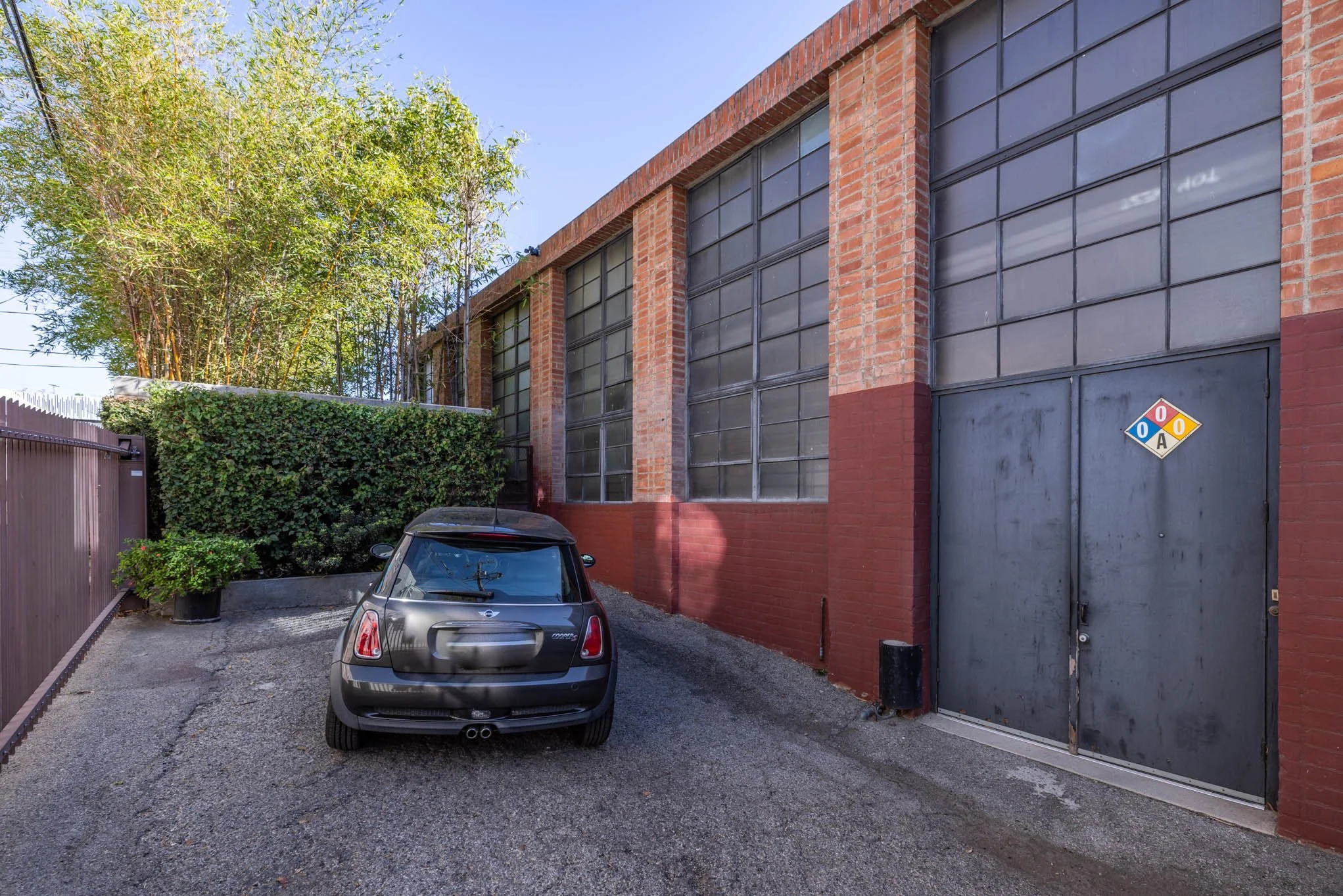 A gray car parked in front of a brick building with large glass windows and a blue double door with a hazard diamond sign.