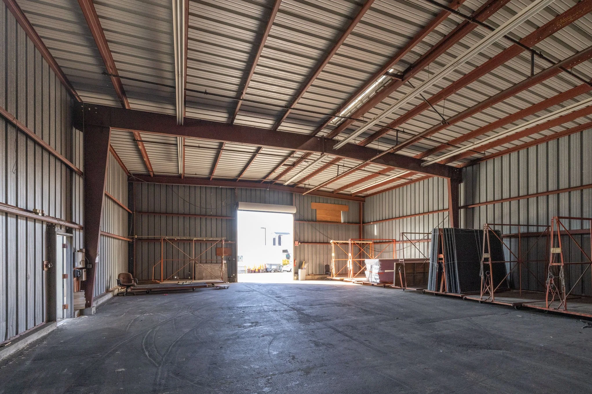 Interior of an empty warehouse with metal walls and ceiling, construction equipment, and an open garage door letting in sunlight.
