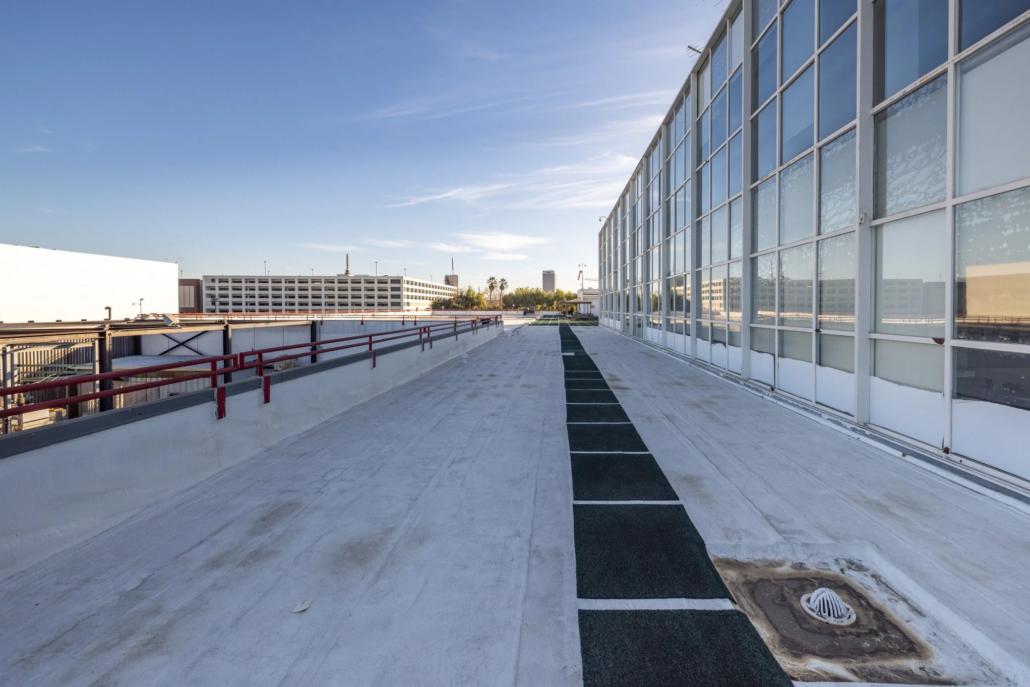 Empty rooftop parking area with black parking strips, red safety railings on the left, and large glass windows on the right under a clear blue sky.