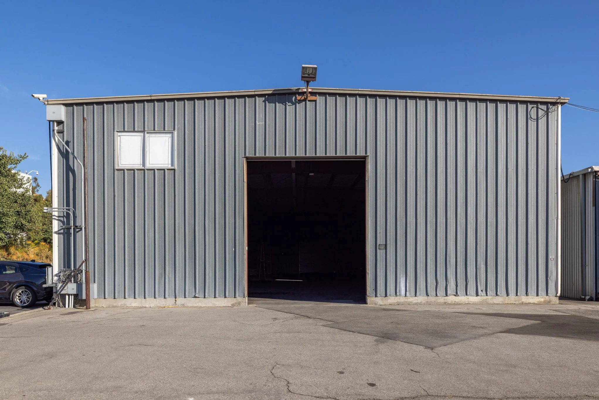 Gray metal warehouse building with large open door, small white window, and parking lot in front.