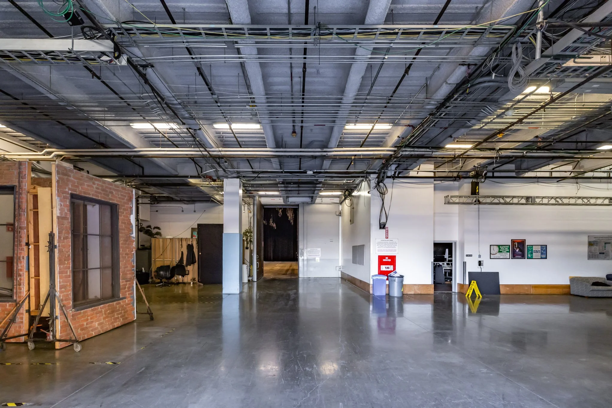 Empty indoor space with visible ceiling pipes and wires, a small brick structure on the left, trash bins, and some chairs and furniture in the background.