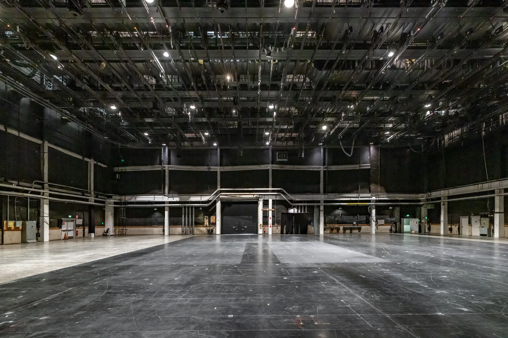 An empty large theater or auditorium with a black stage and high ceiling, with lighting and rigging equipment hanging above.