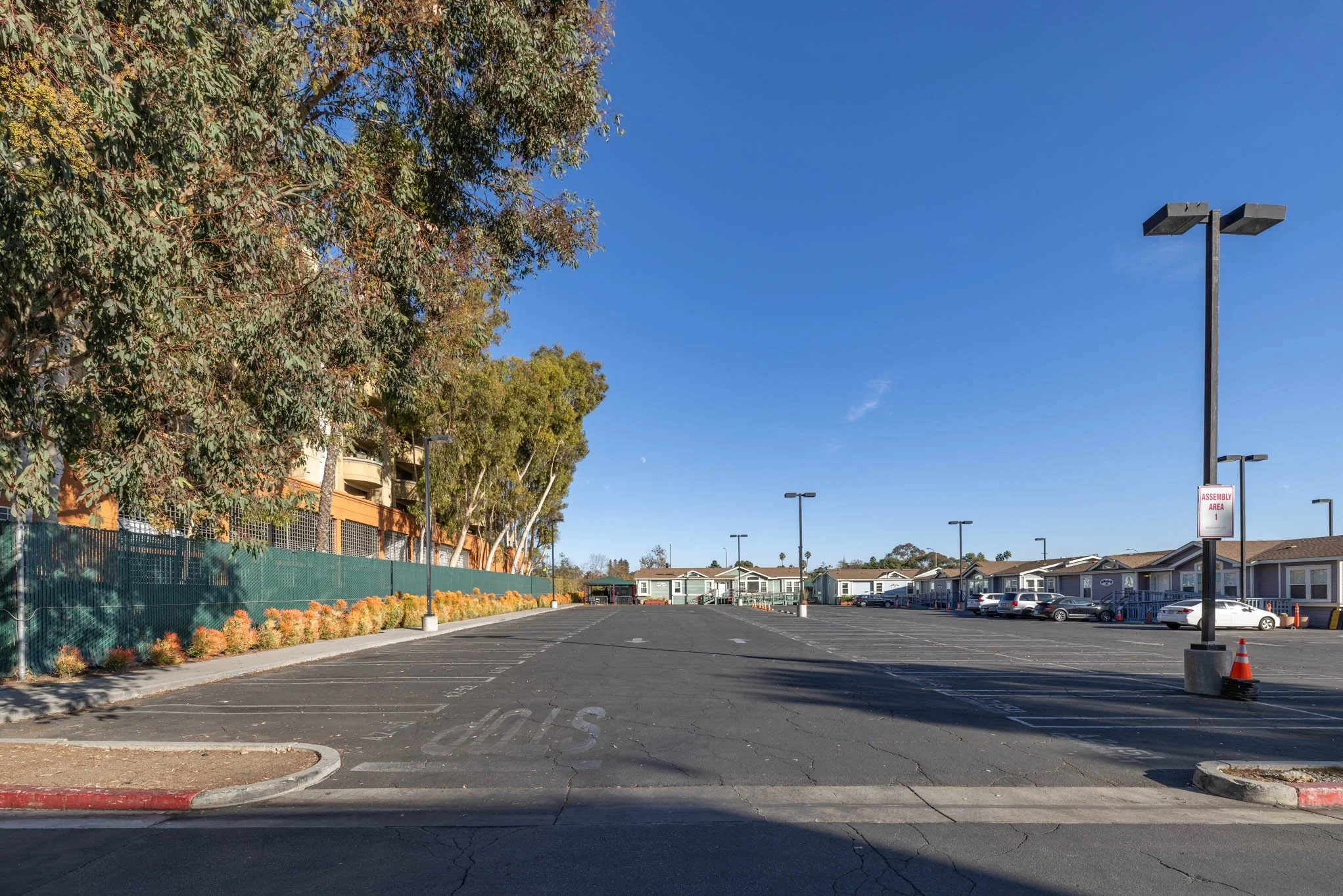 Empty parking lot with residential units in the background under a clear blue sky, trees on the left, parking space lines and a sign that reads 'Assembly Area' on a lamppost.