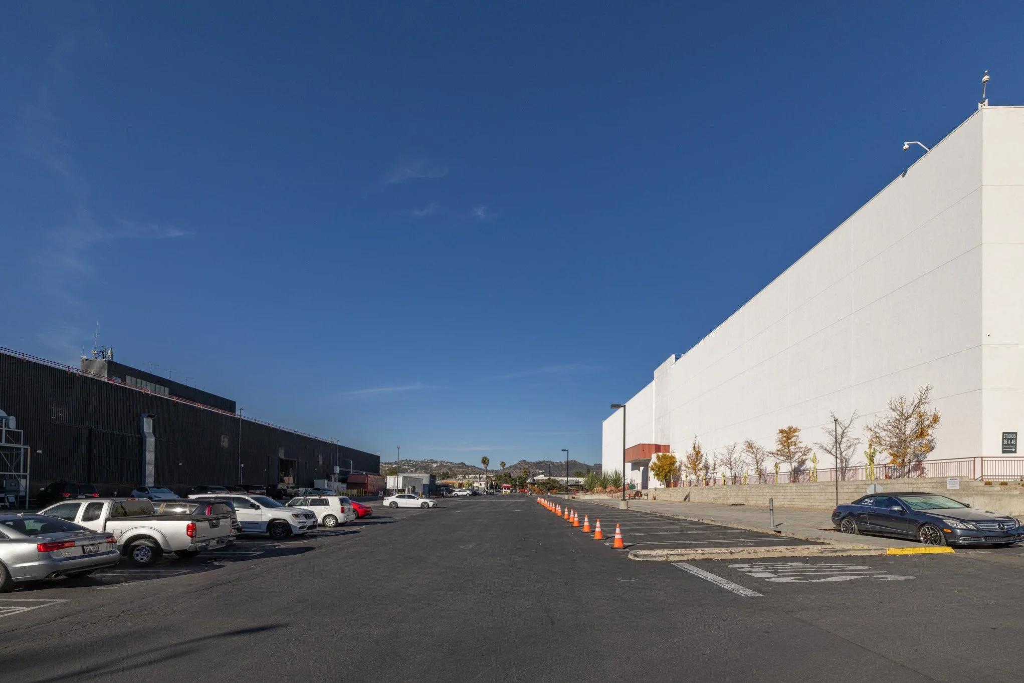 Large outdoor parking lot with several cars parked, orange traffic cones creating a barrier, and a large white building on the right side of the image under a clear blue sky.